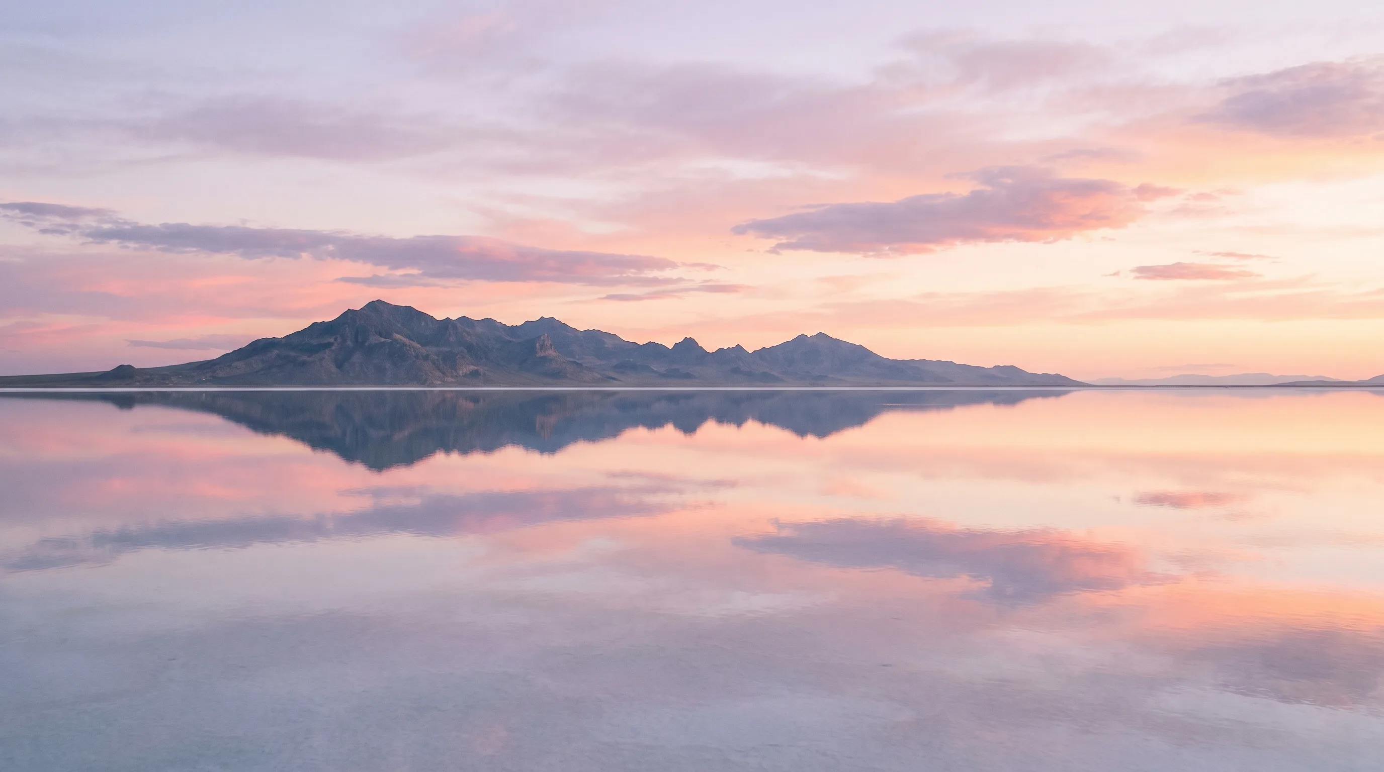 The Bonneville Salt Flats at sunrise, a thin sheet of standing water turning the white salt into a perfect mirror reflecting distant mountains and a pastel sky.