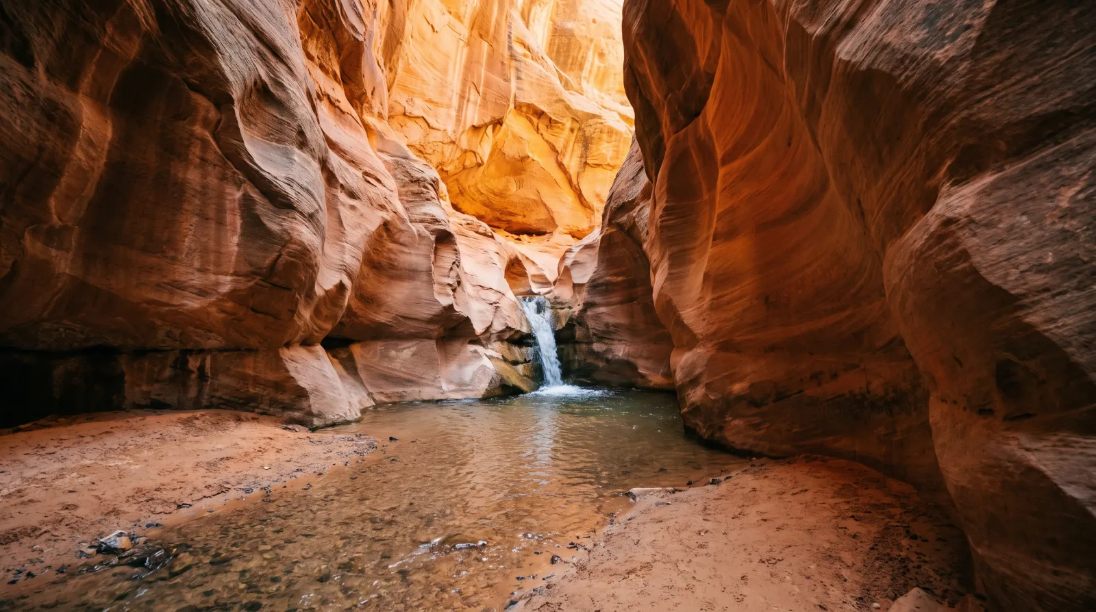 A hiker wading up the glowing orange sandstone slot canyon toward Kanarraville Falls in southern Utah, with the waterfall and aluminum stairs visible ahead.