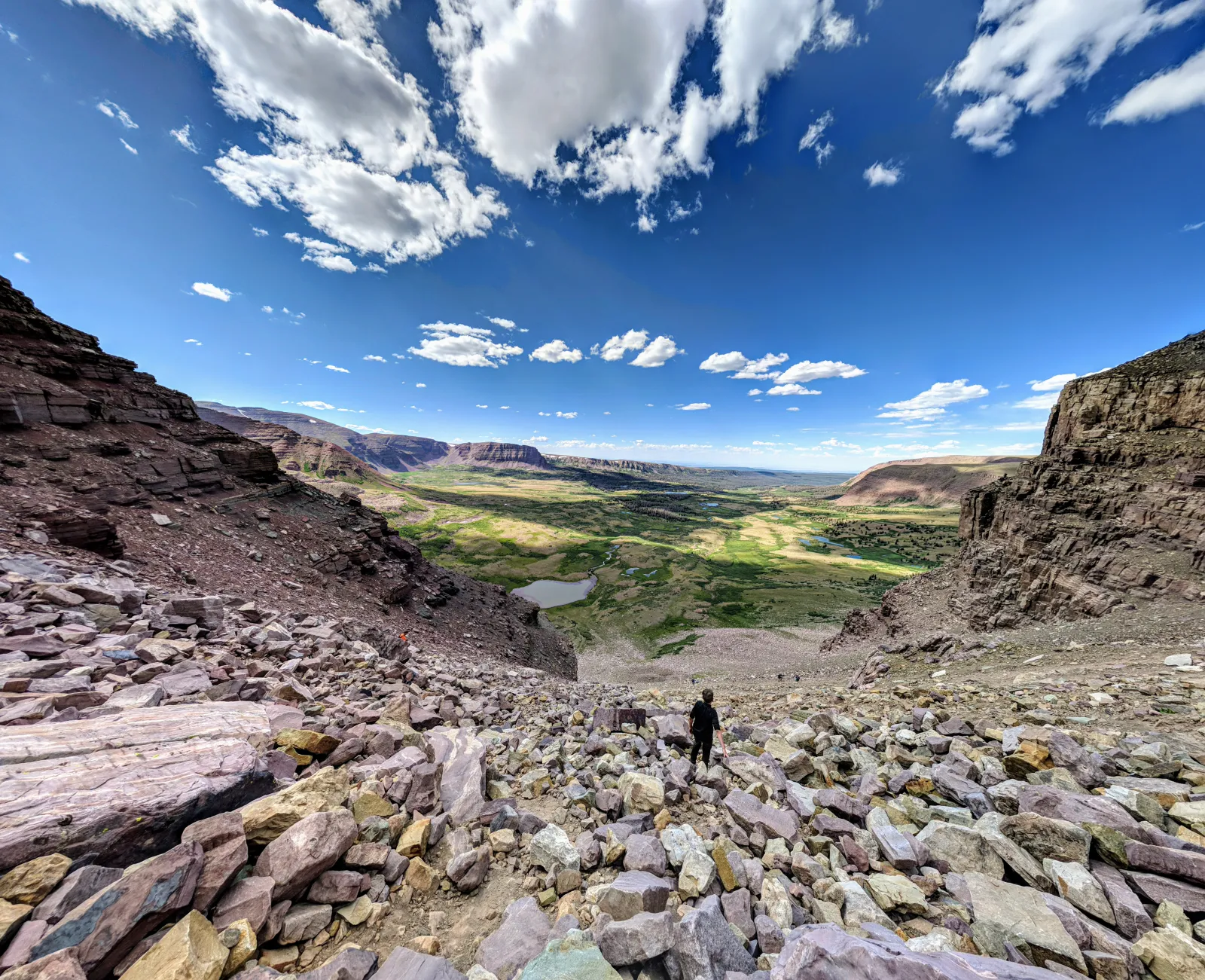 A lone hiker descending a boulder-strewn talus slope high on Kings Peak, looking out over the vast green Henry's Fork basin dotted with alpine lakes under a bright blue sky with scattered clouds, in the High Uintas Wilderness of northeastern Utah.