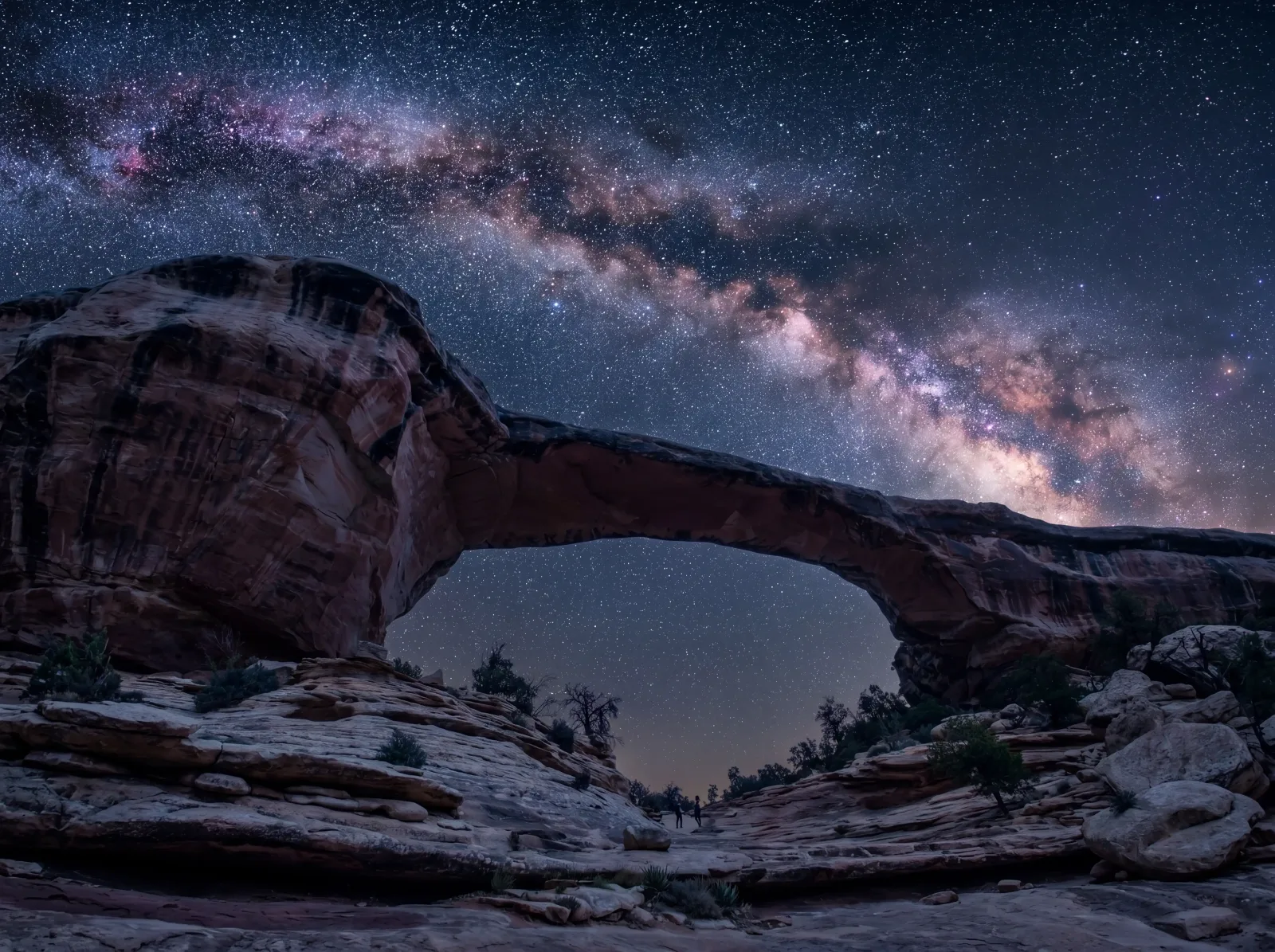 The Milky Way arching over a red rock natural bridge under a deep, star-filled desert sky.