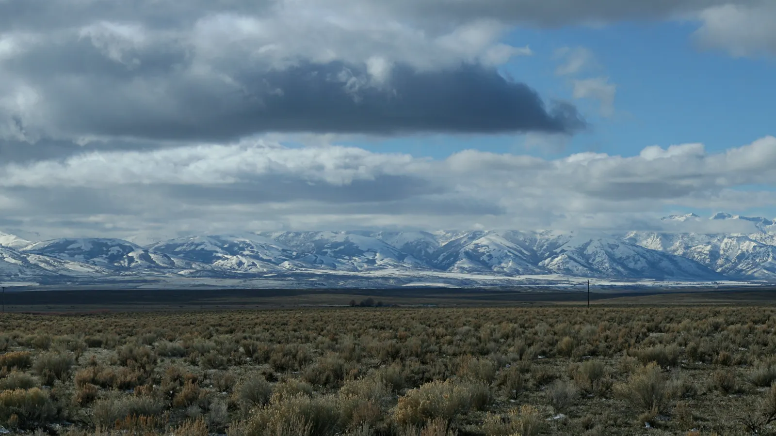 The wide, empty Great Basin Desert — a flat sagebrush plain running to a distant snow-dusted mountain range under a big cloud-filled sky — the kind of vast, remote high-desert basin that surrounds Nancy Holt's Sun Tunnels in far northwestern Utah.