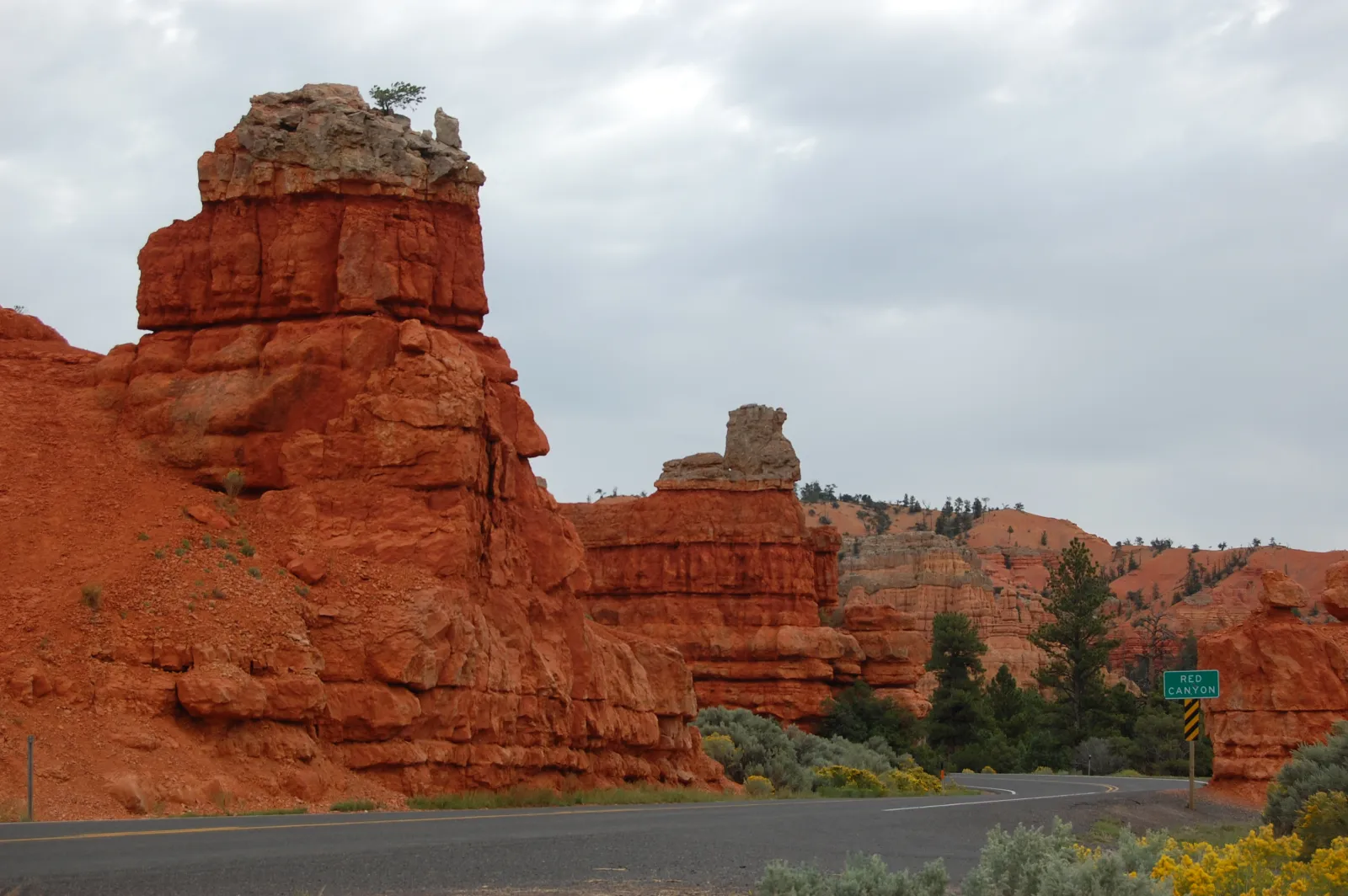 A highway curving through red rock hoodoos past a green "Red Canyon" road sign on Utah's Scenic Byway 12.